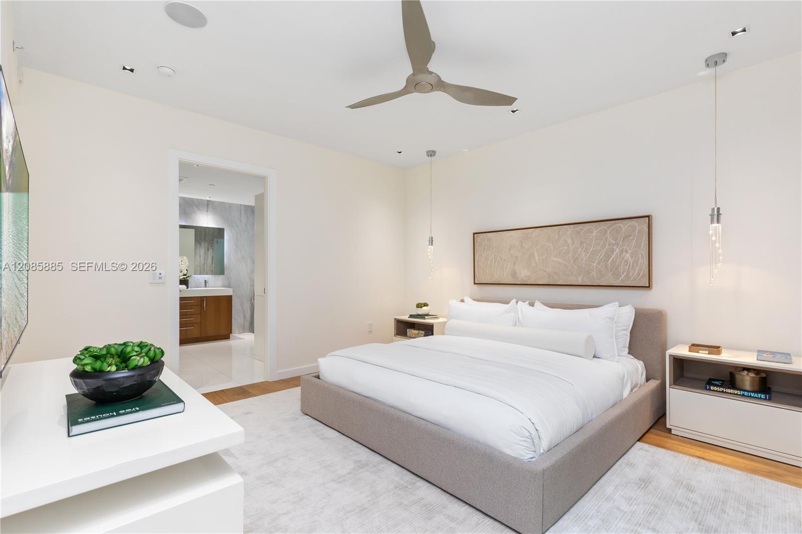 Staged bedroom with a large white bed, upholstered headboard, ceiling fan, and bathroom doorway.