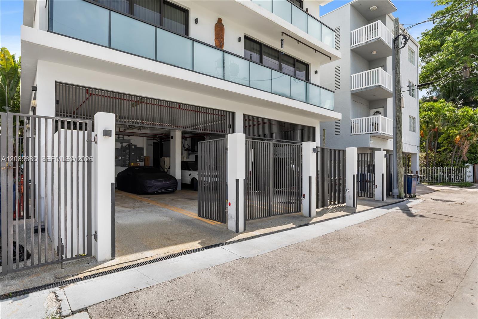 Modern white condominium building with glass balconies and gated ground-floor parking.