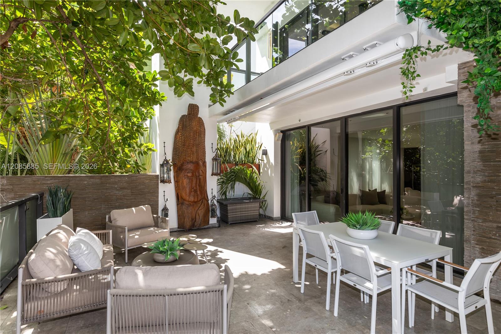 Furnished terrace with white dining set, cushioned lounge seating, leafy canopy, and glass doors.