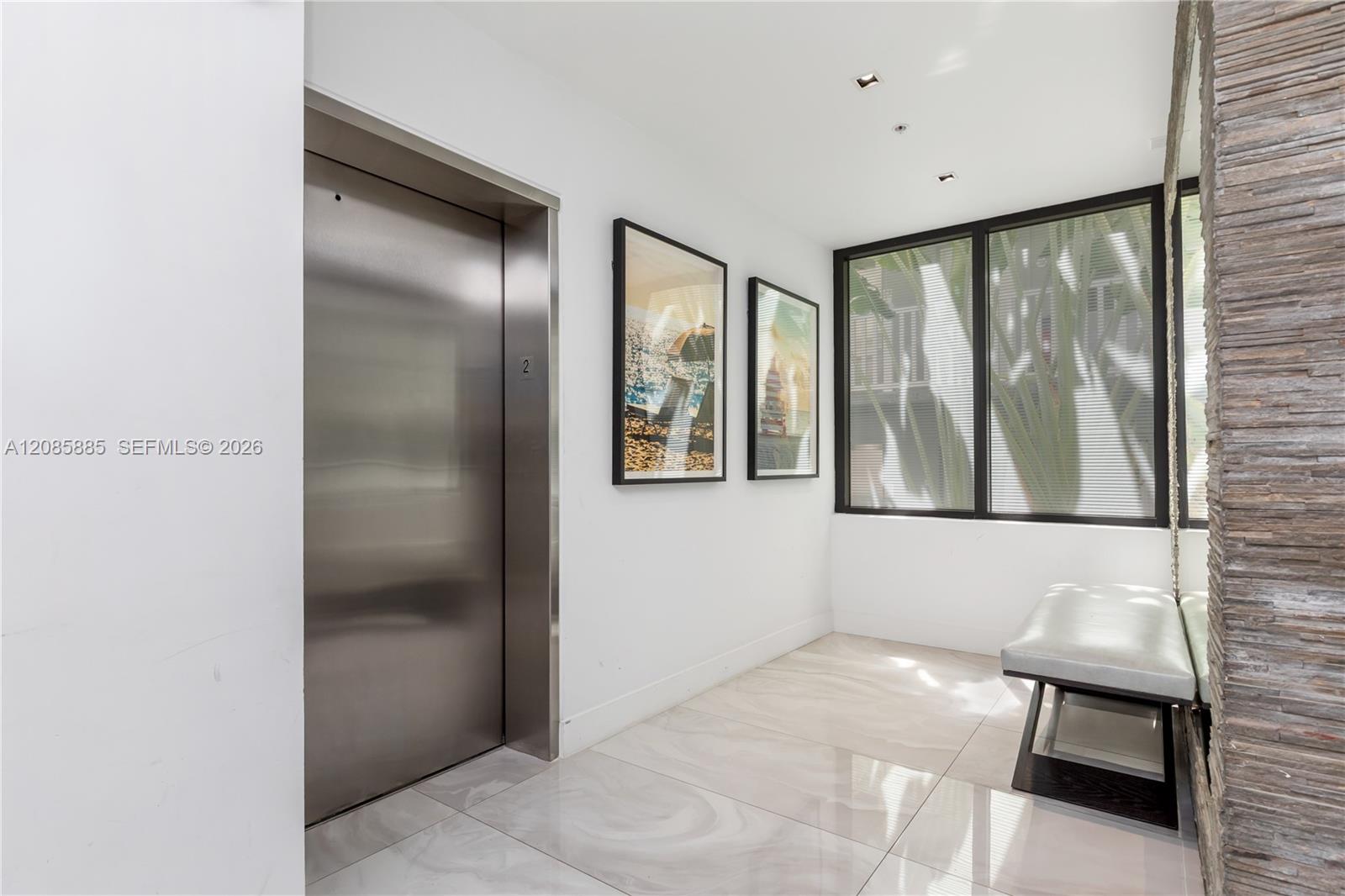 Modern lobby with a stainless-steel elevator, framed artwork, windows, and a bench beside a stone wall.