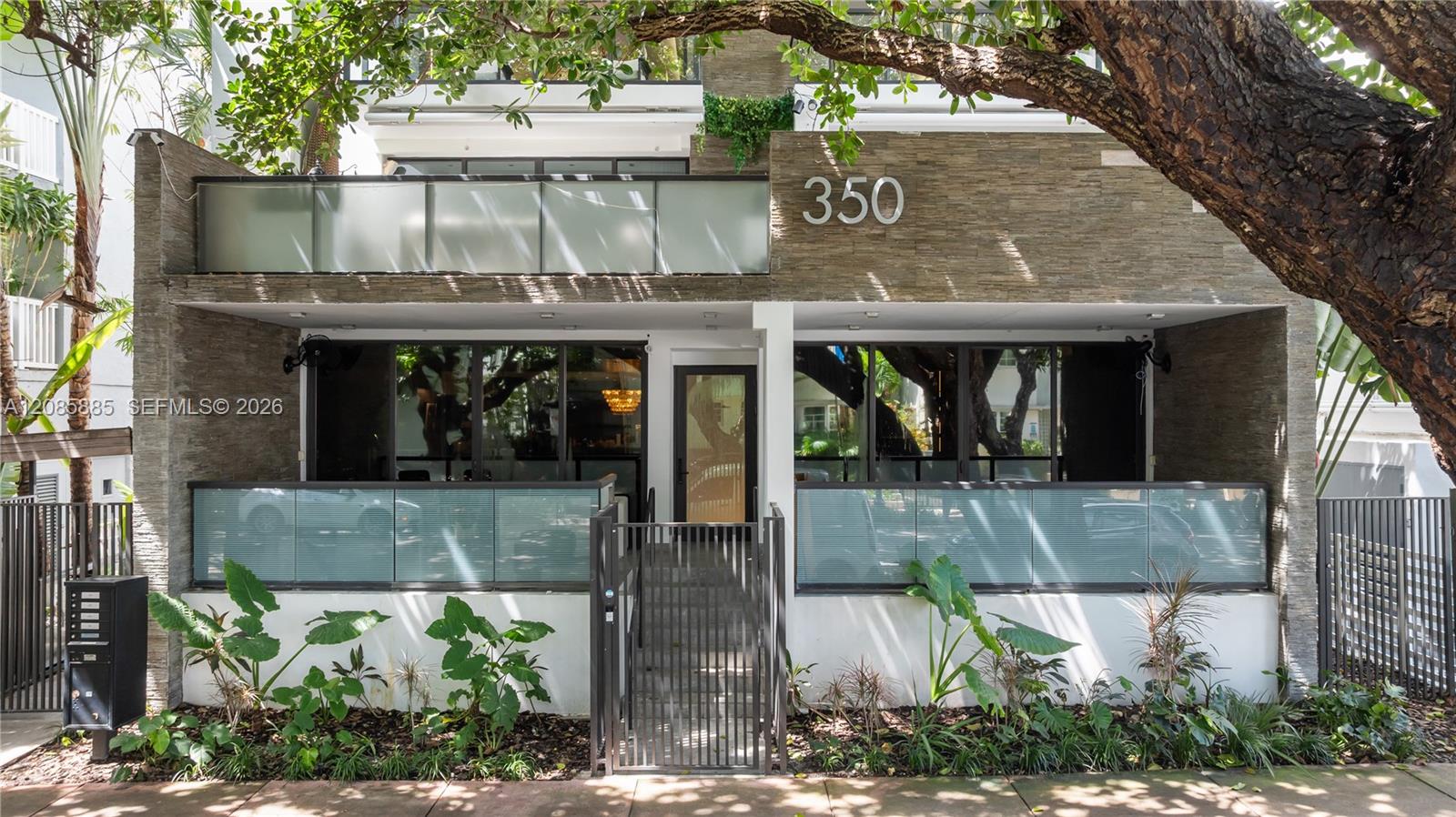 Modern two-story residence with stone facade, glass balconies, gated entry and a large shade tree.