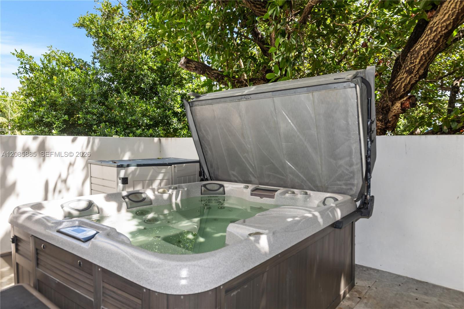 Outdoor hot tub with its cover raised on a shaded patio surrounded by leafy trees.