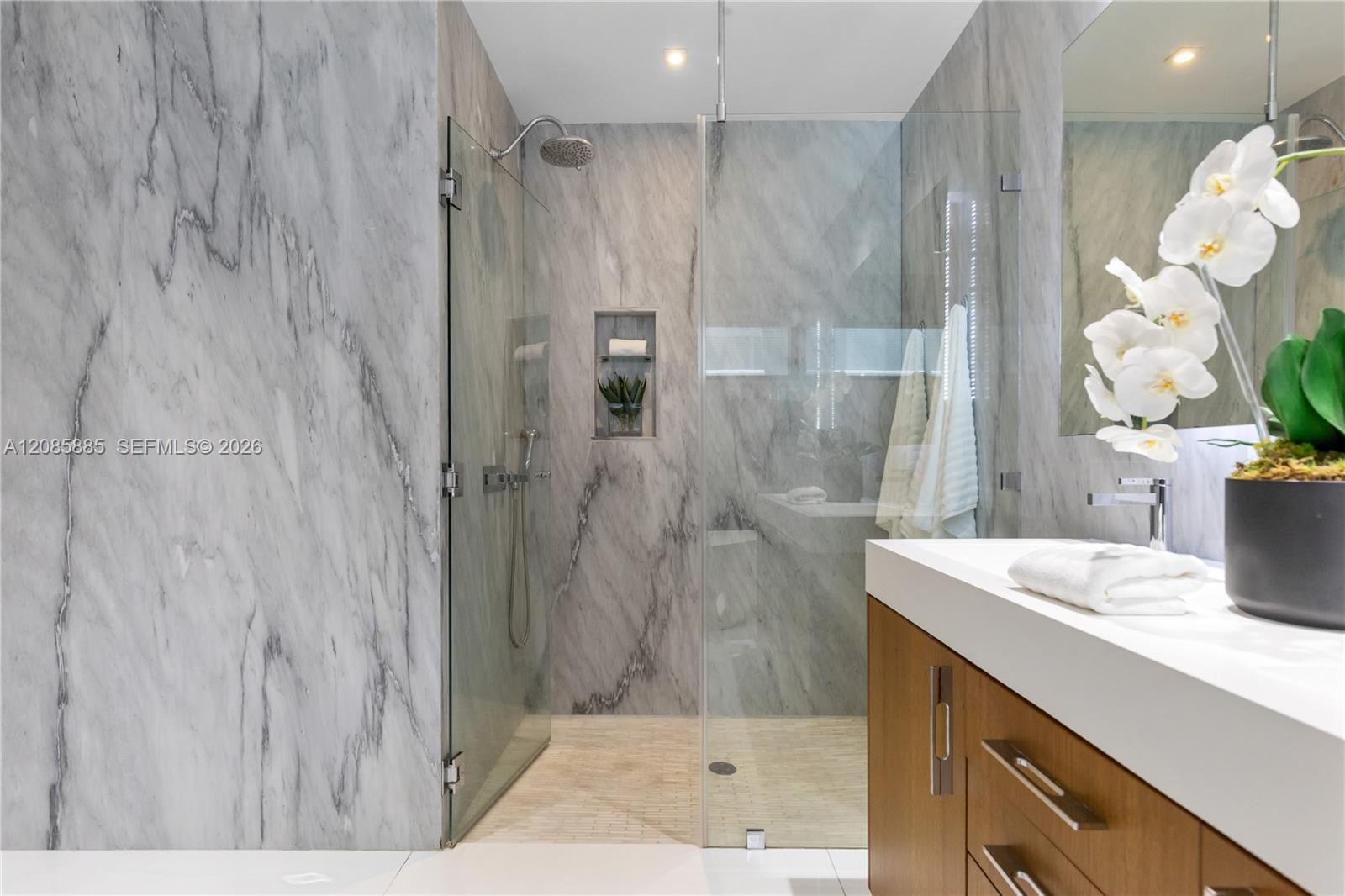 Marble bathroom with a glass walk-in shower and wood vanity topped by a white orchid.