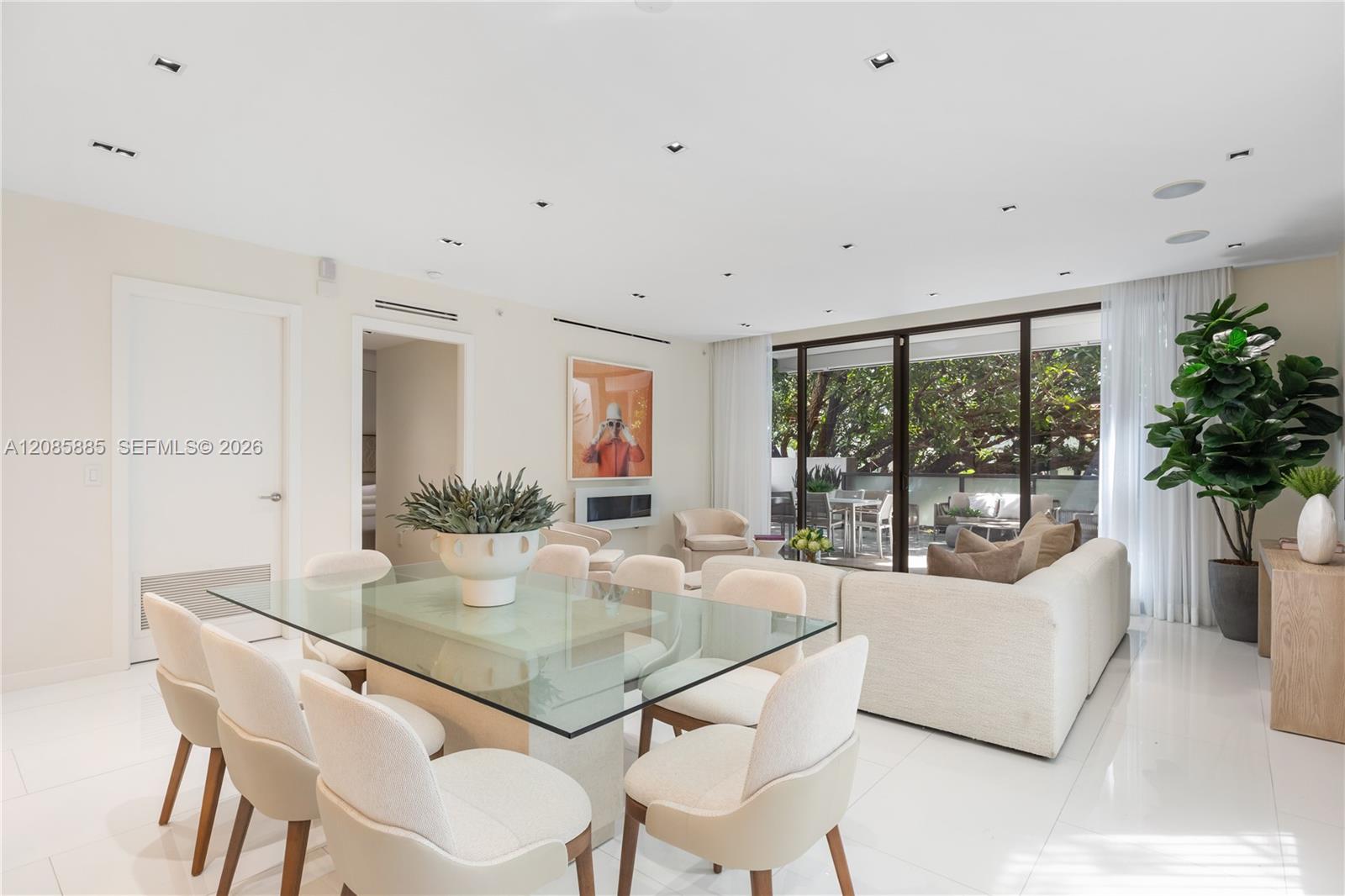 Open-plan dining and living room with a glass dining table, cream furnishings, and leafy patio views.