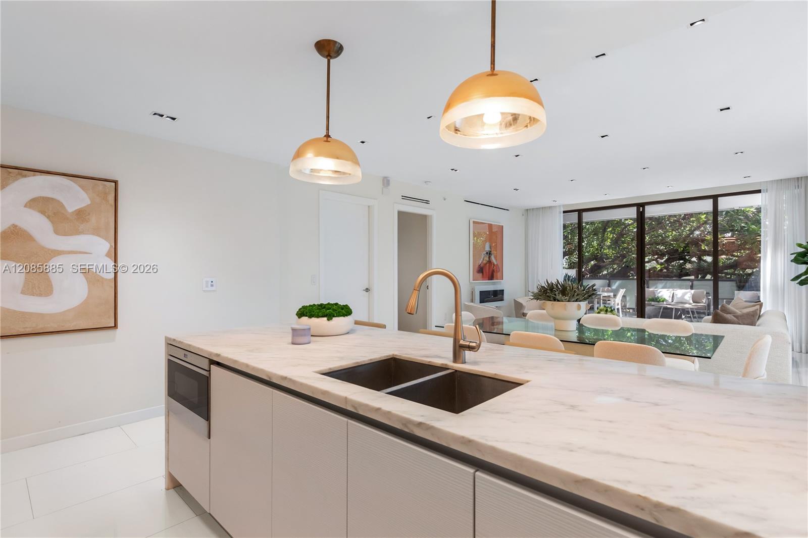 Open-plan kitchen with a marble island and double sink, overlooking a furnished living area and leafy patio.