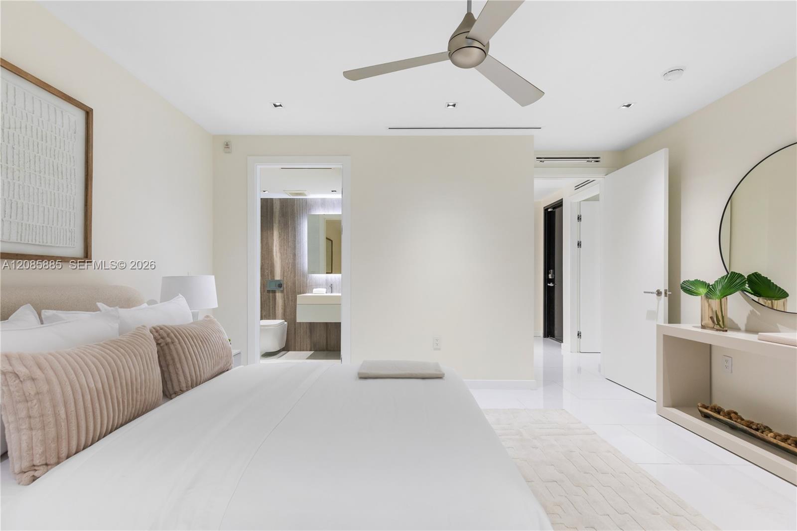Staged bedroom with a white bed, blush pillows, ceiling fan, and open doorway to an ensuite bathroom.