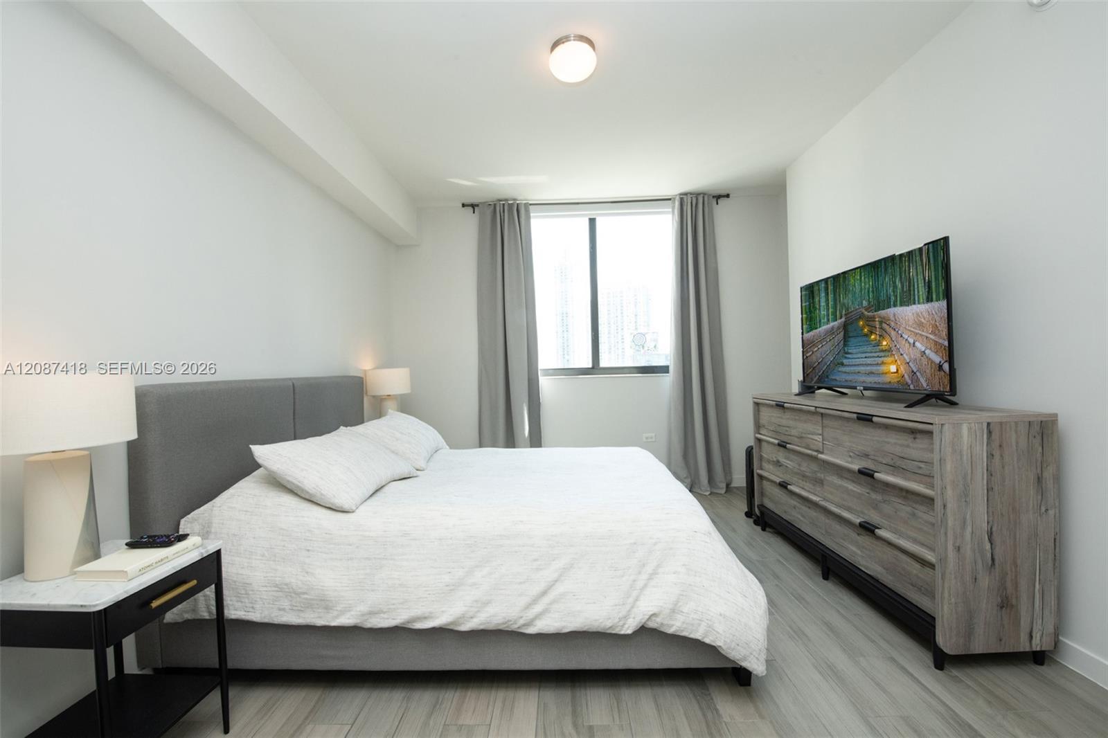 Furnished bedroom with a large white bed, gray headboard, and television on a wood-look dresser.