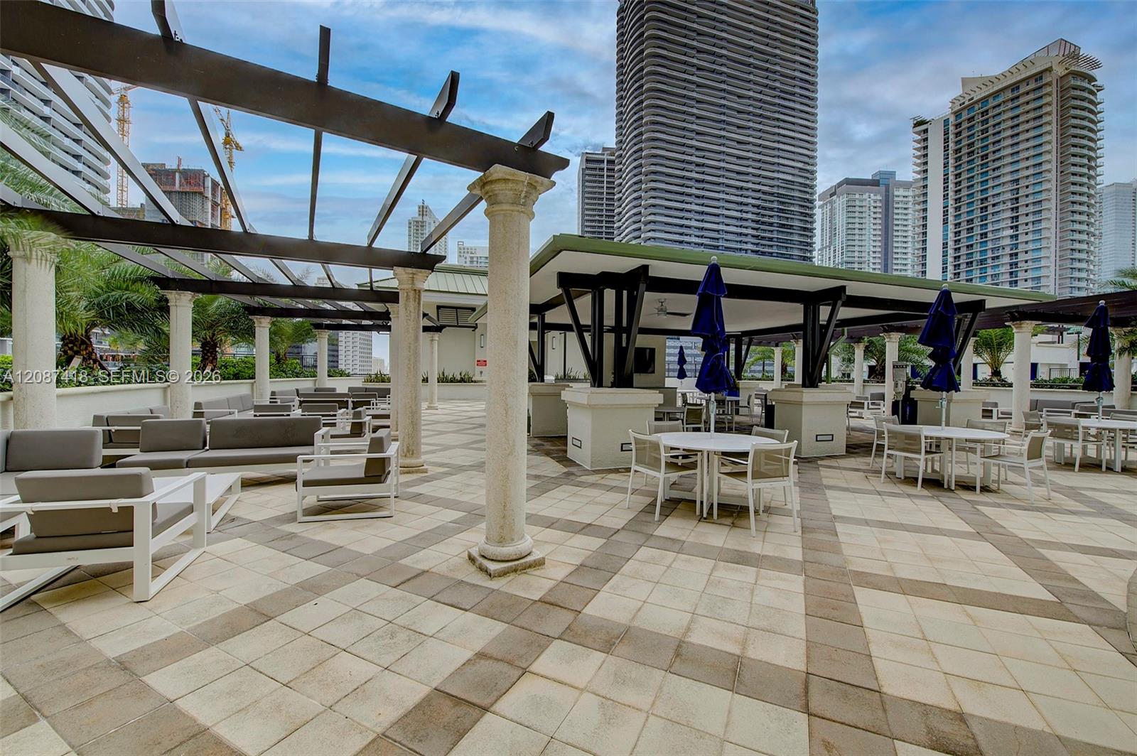 Condominium terrace with pergola, tiled paving, lounge seating, and dining tables against a high-rise skyline.