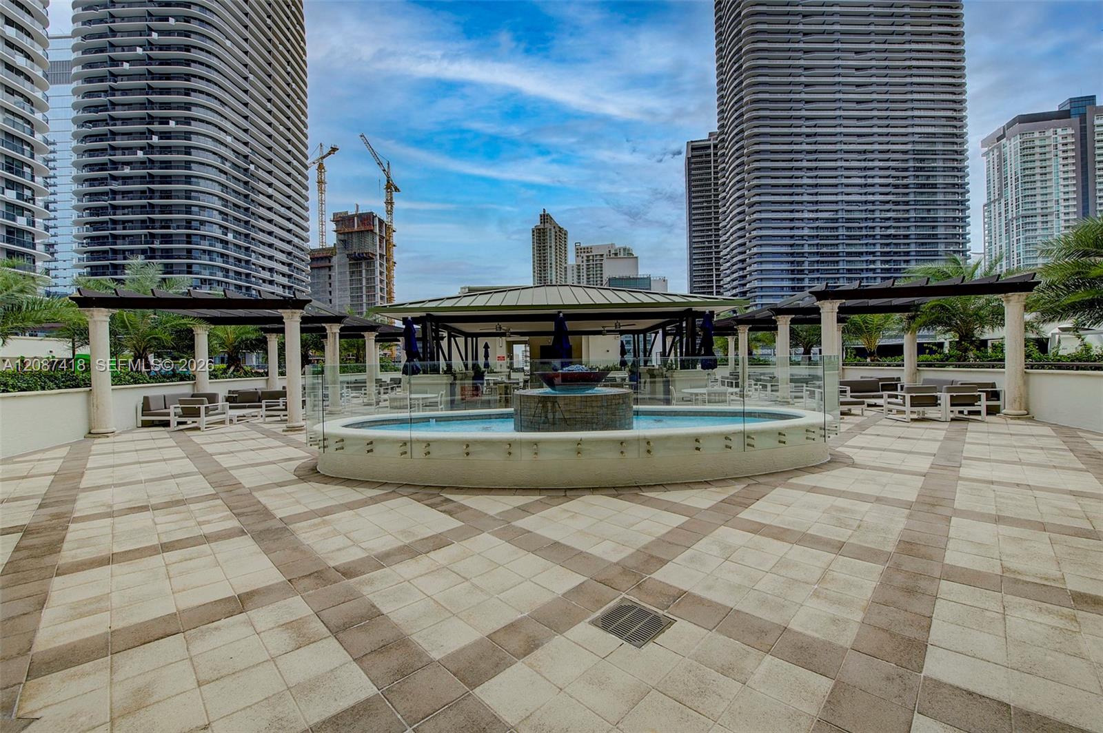 Condominium pool deck with a circular fountain pool, pergola-covered seating, and high-rise buildings.