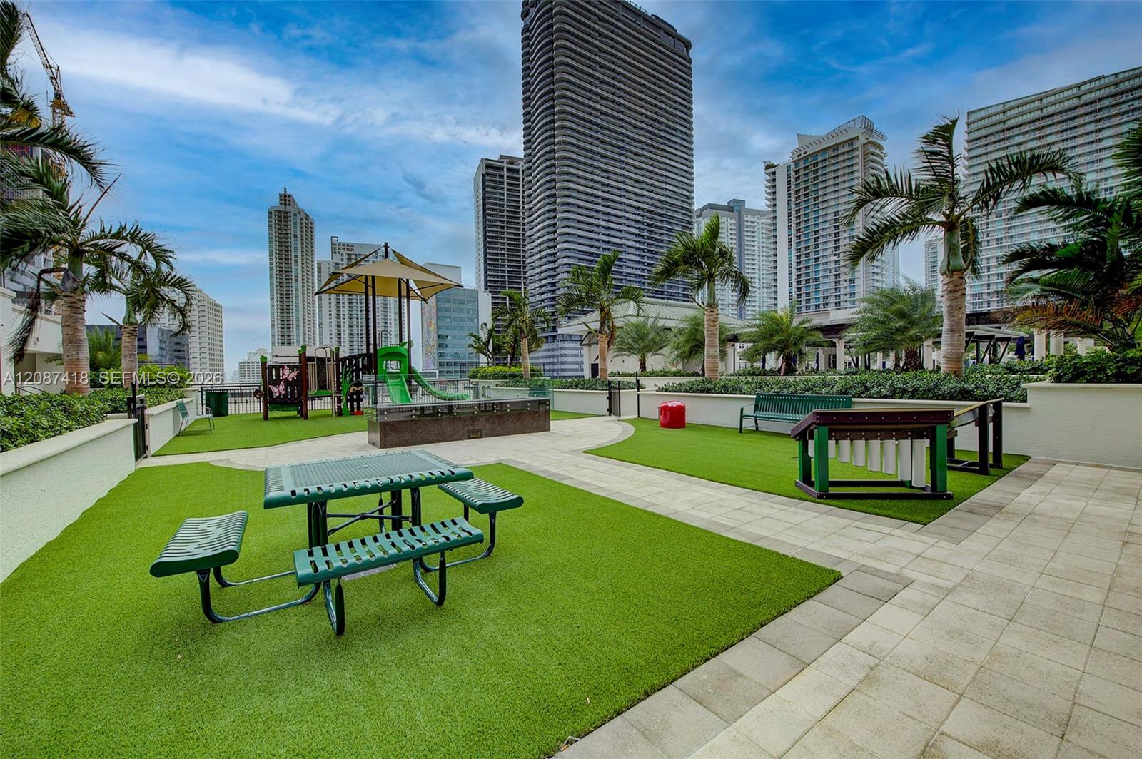 Outdoor recreation terrace with turf, picnic tables, a playground, and palm trees against a high-rise skyline.