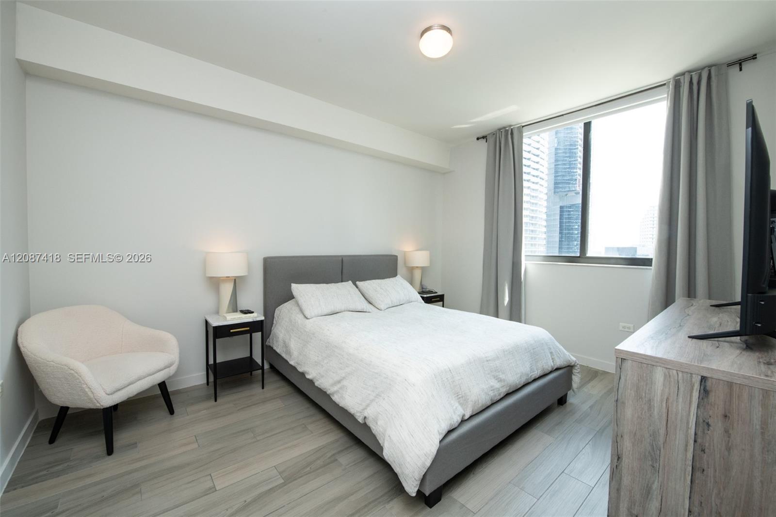 Furnished bedroom with a gray bed, cream chair, and window overlooking downtown high-rises.