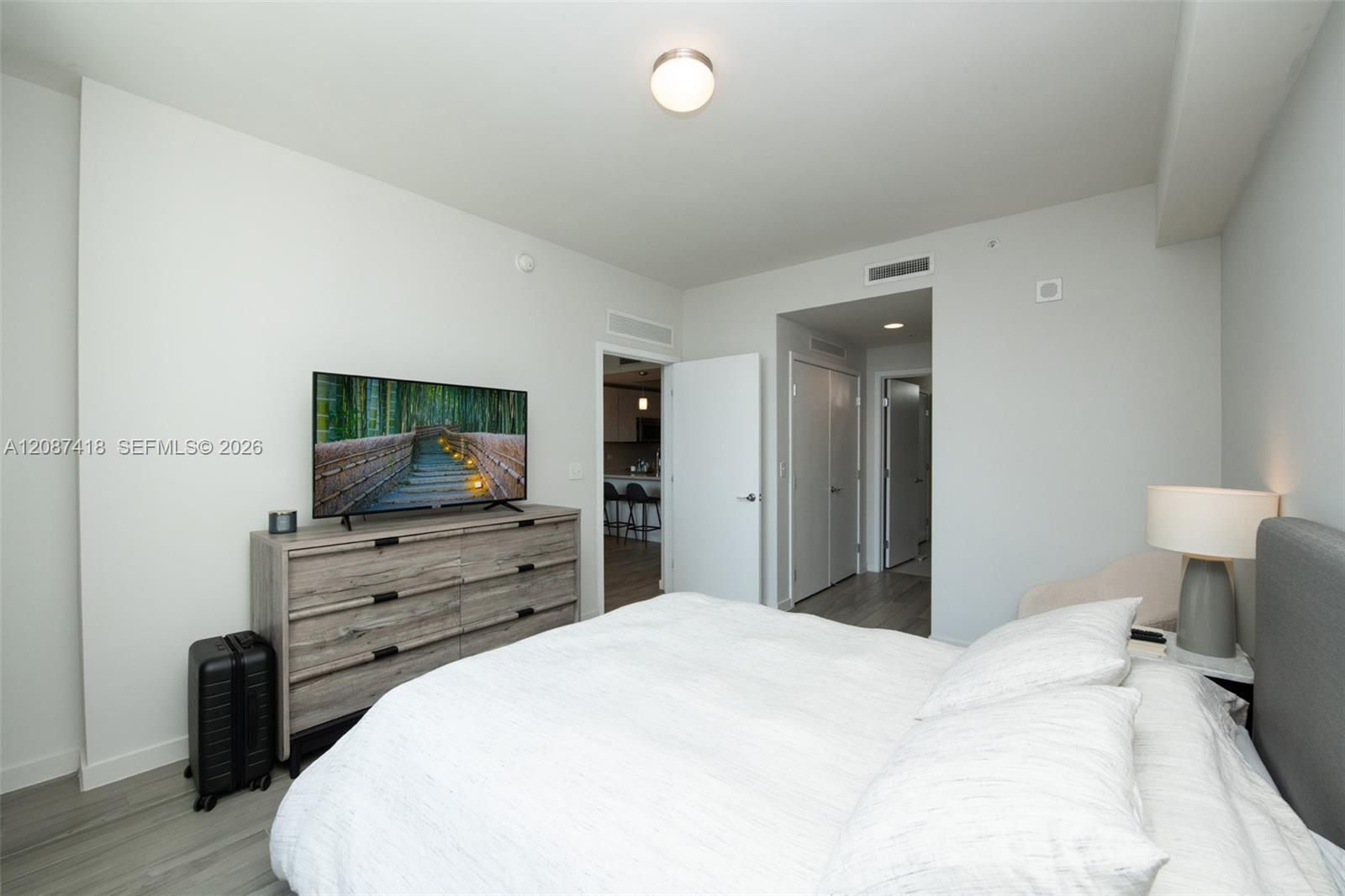 Furnished bedroom with a white bed, gray headboard, and wood-grain dresser topped by a television.