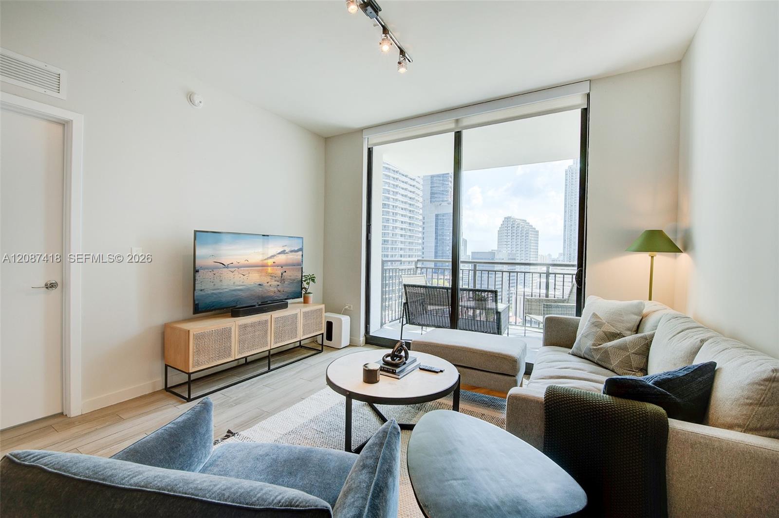 Furnished living room with sofas, a balcony, and views of nearby high-rise buildings.