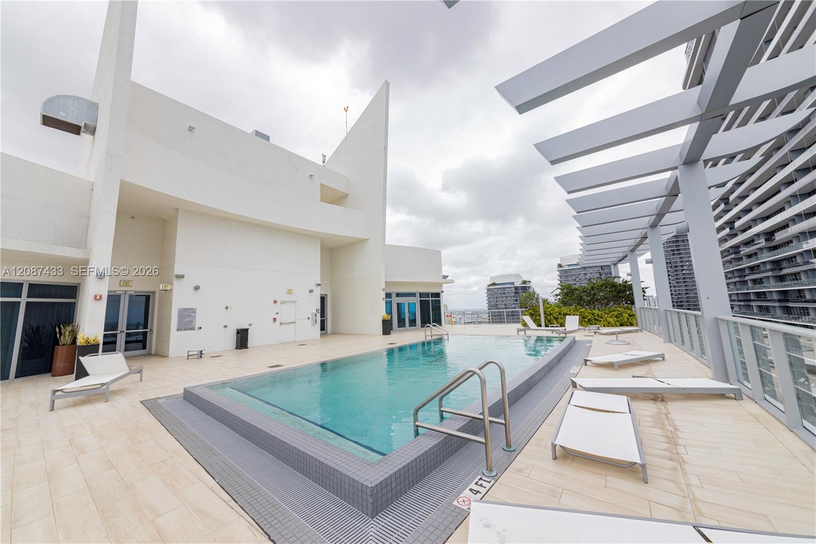 Rooftop pool bordered by white loungers and a modern pergola, with neighboring high-rises beyond.
