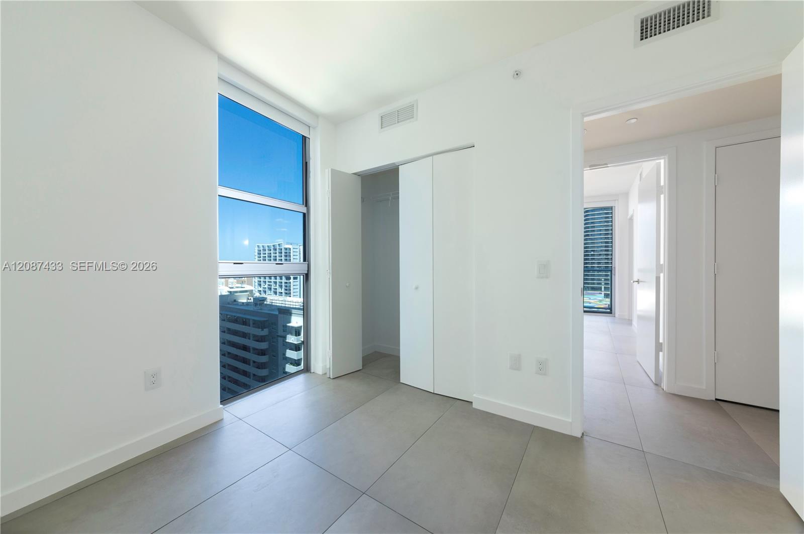 Vacant condo bedroom with a large window, sliding closet, and views of nearby buildings.
