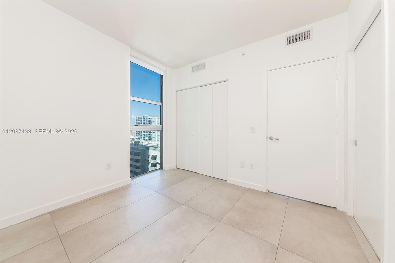 Vacant room with tiled floor, closet doors, and a window overlooking nearby buildings.