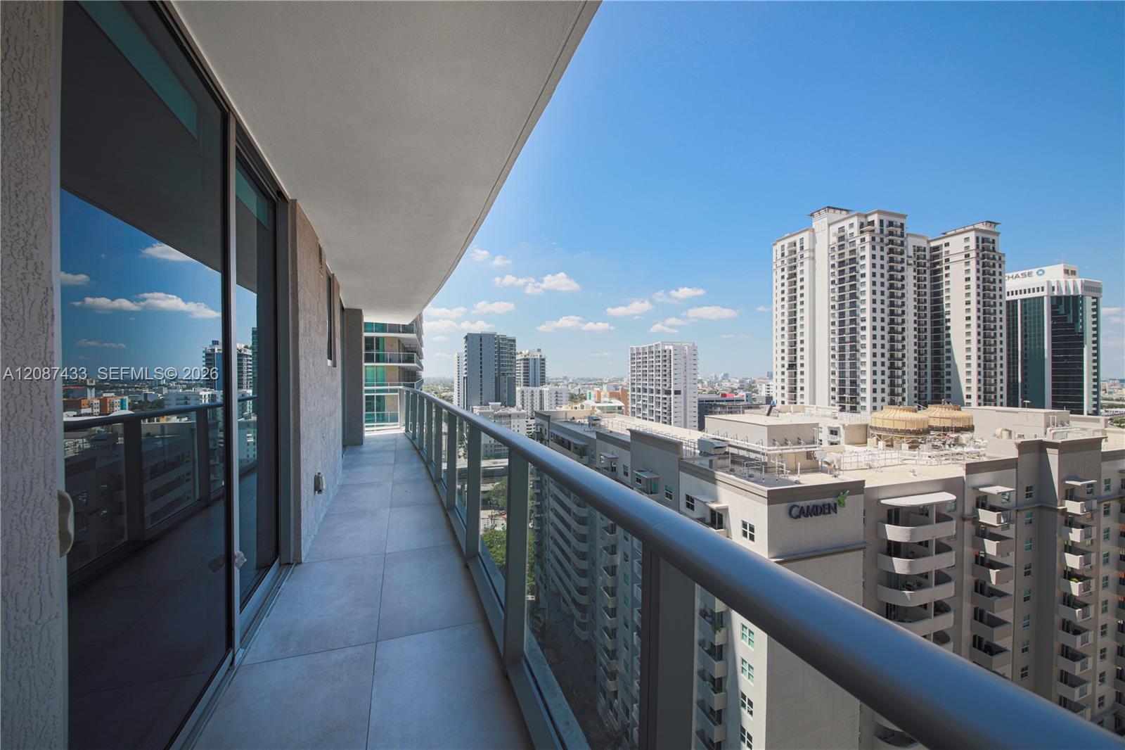 High-rise condo balcony with a glass railing overlooking downtown buildings under a bright blue sky.