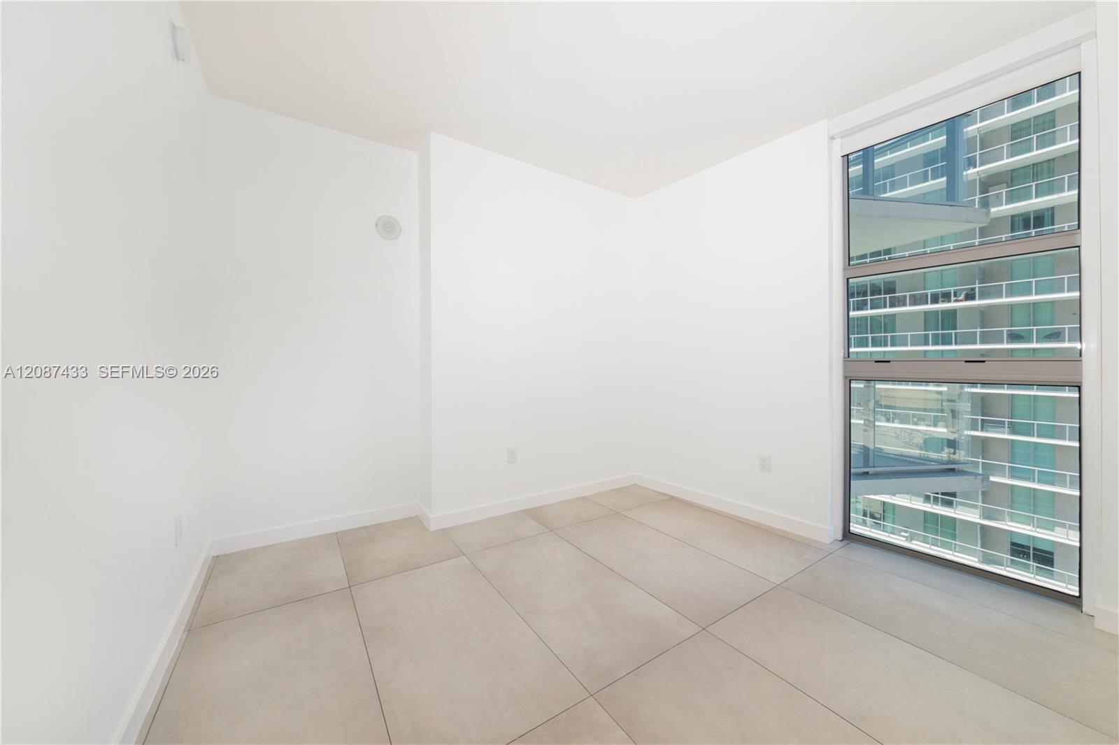 Vacant condominium room with large tile flooring and a tall window facing a neighboring high-rise.