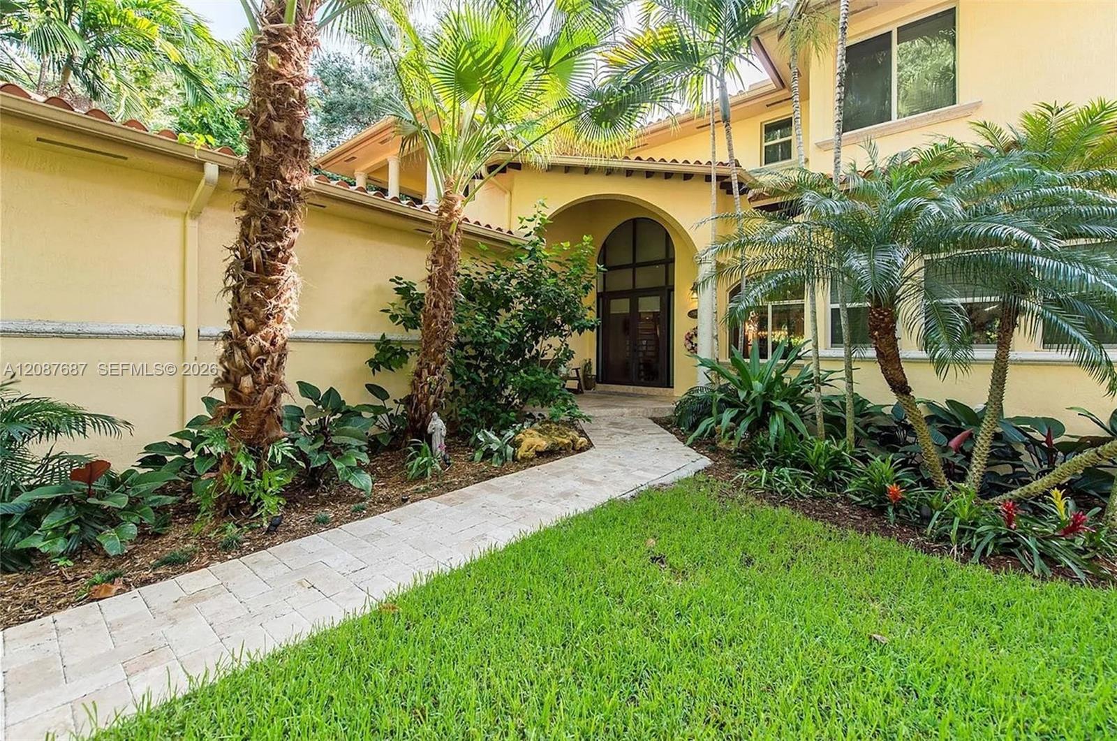 Yellow stucco home entrance framed by palm trees and tropical landscaping, with a curved stone walkway.