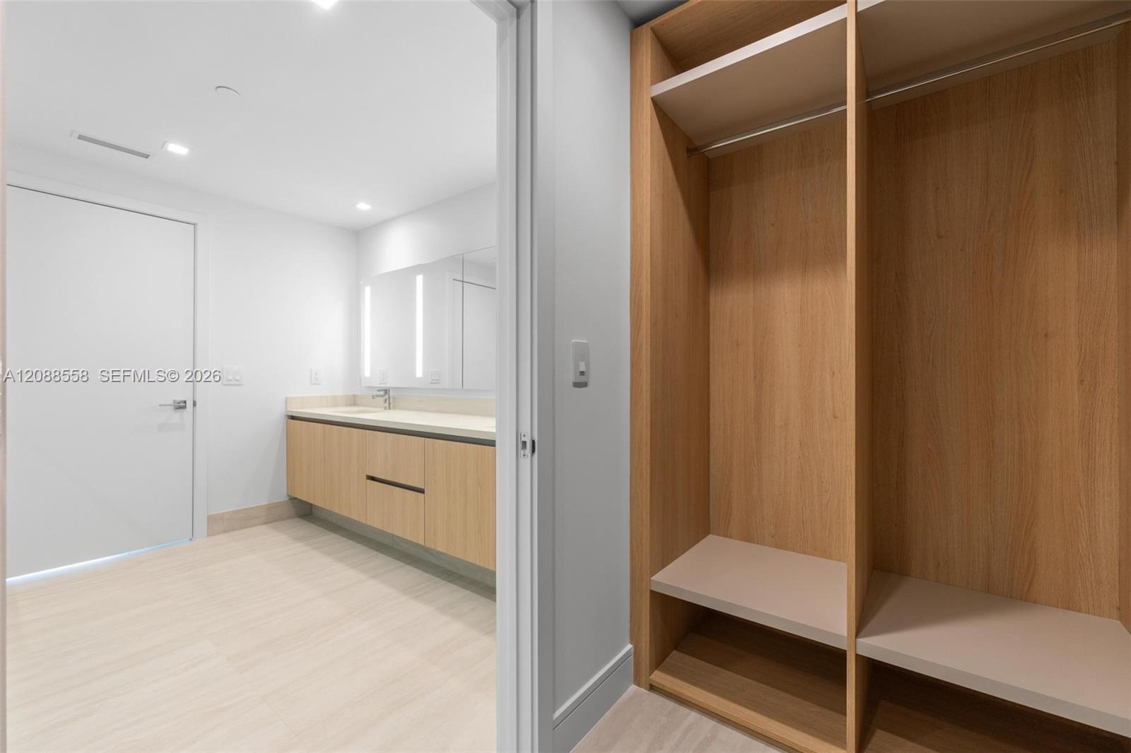 Walk-in closet with light wood shelving beside a bright bathroom vanity.
