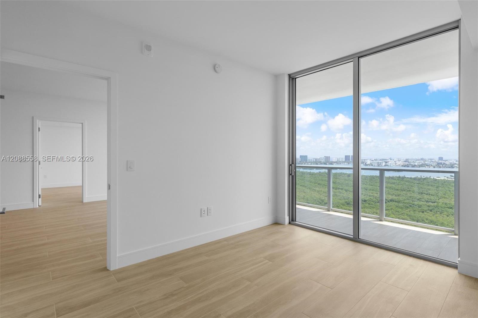 Vacant condo room with light wood-look floors and balcony views of wetlands, water, and the skyline.