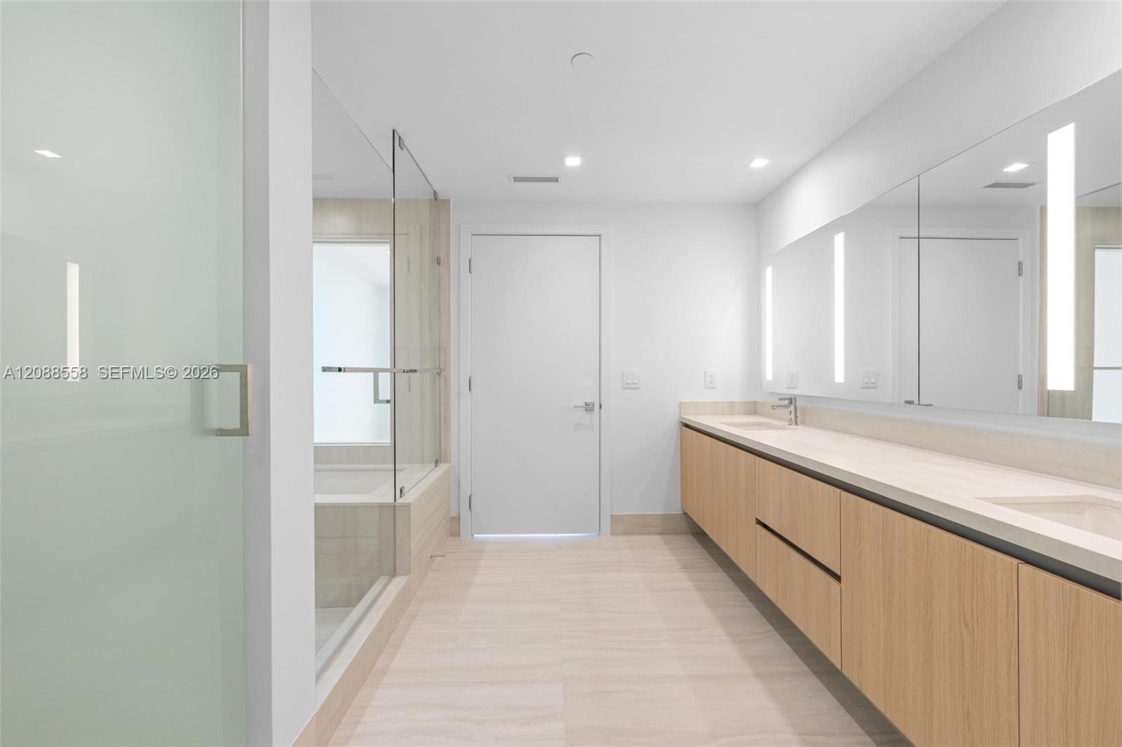 Modern bathroom with a long light-wood vanity, pale stone counter, large mirrors, and glass-enclosed tub.