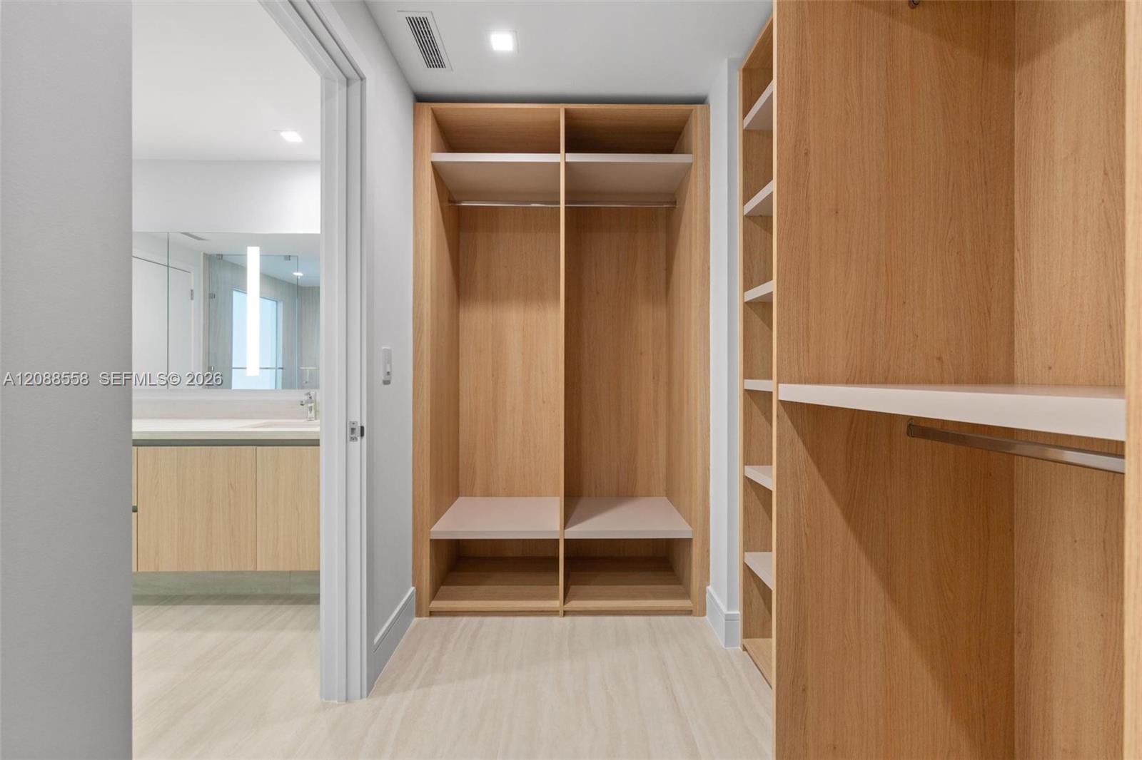 Walk-in closet with light oak built-in wardrobes and a bathroom vanity visible through the doorway.
