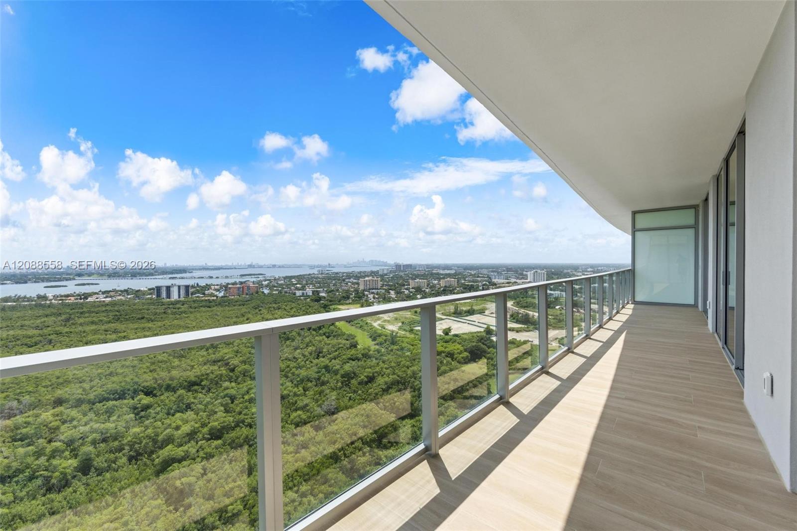 High-rise balcony with glass railing overlooking greenery, water, and distant buildings.