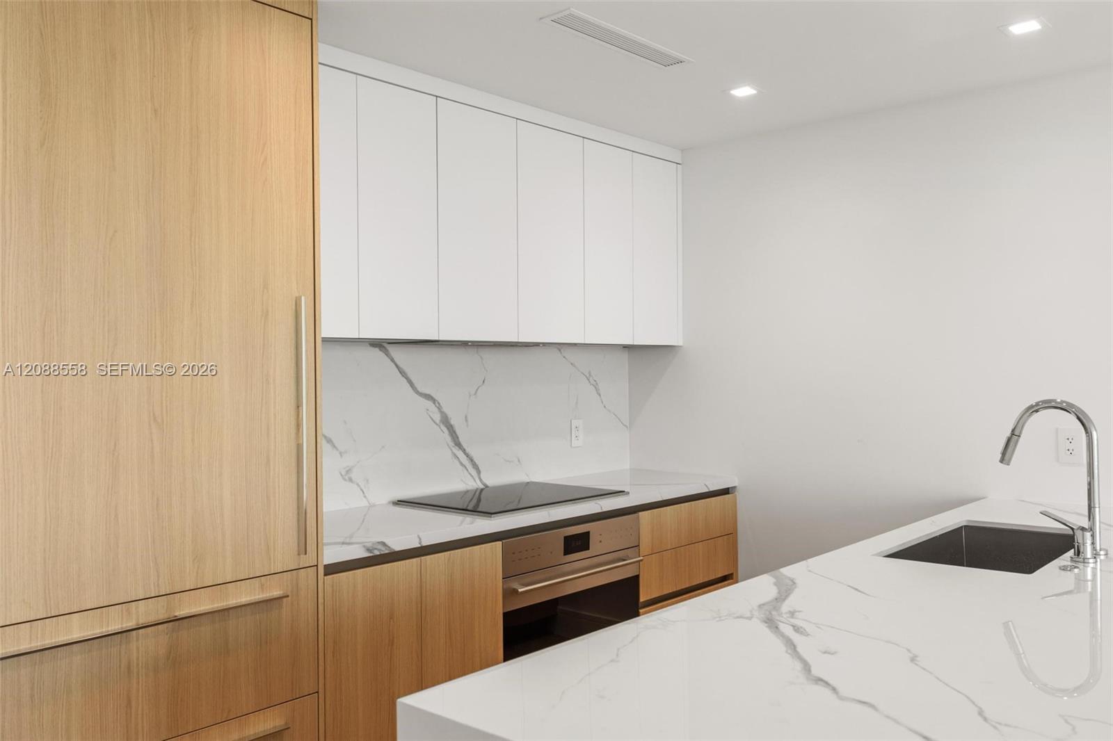 Modern kitchen with light wood cabinets, marble surfaces, built-in cooktop, and island sink.