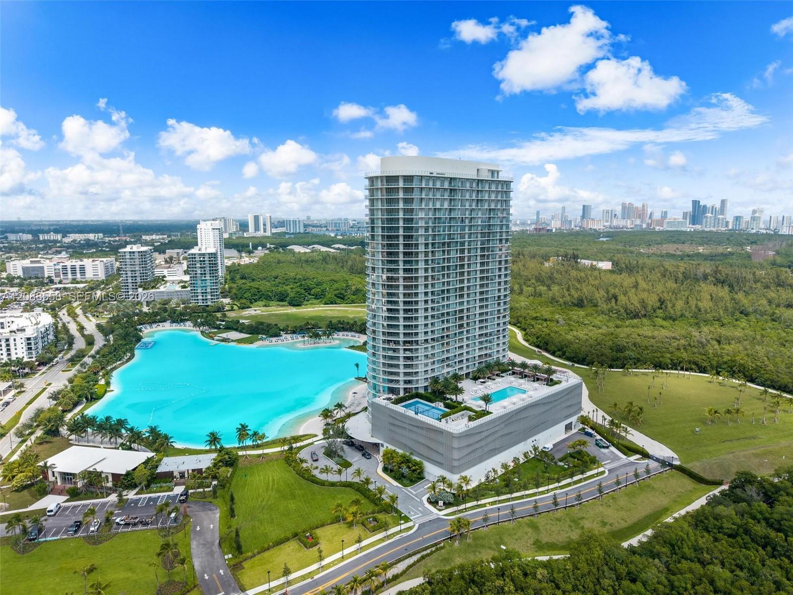 Aerial view of a glass high-rise beside a turquoise lagoon pool, with Miami’s skyline in the distance.