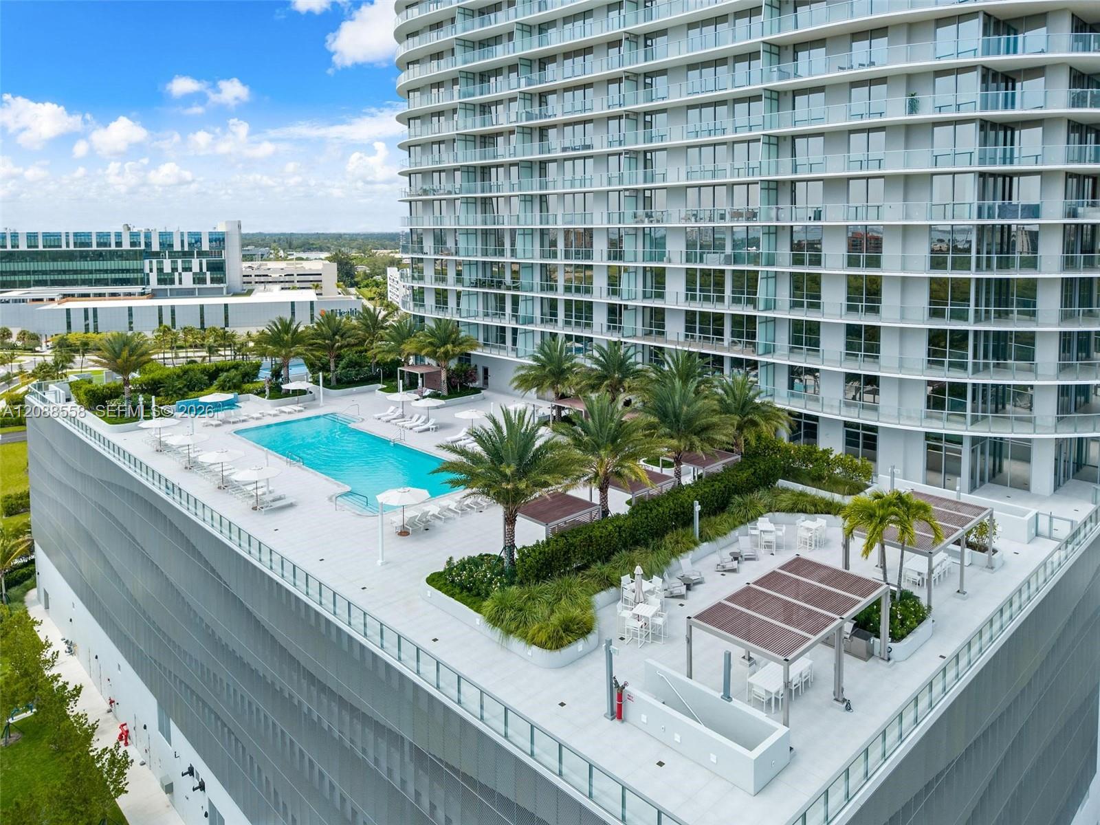 Modern condominium tower beside a rooftop pool deck with palm trees and shaded dining areas.