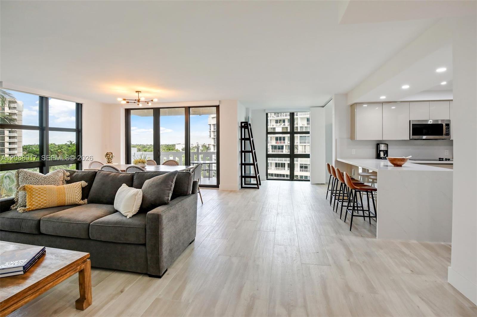 Open-plan condo living room and kitchen with large windows, gray sectional, and views of neighboring buildings.