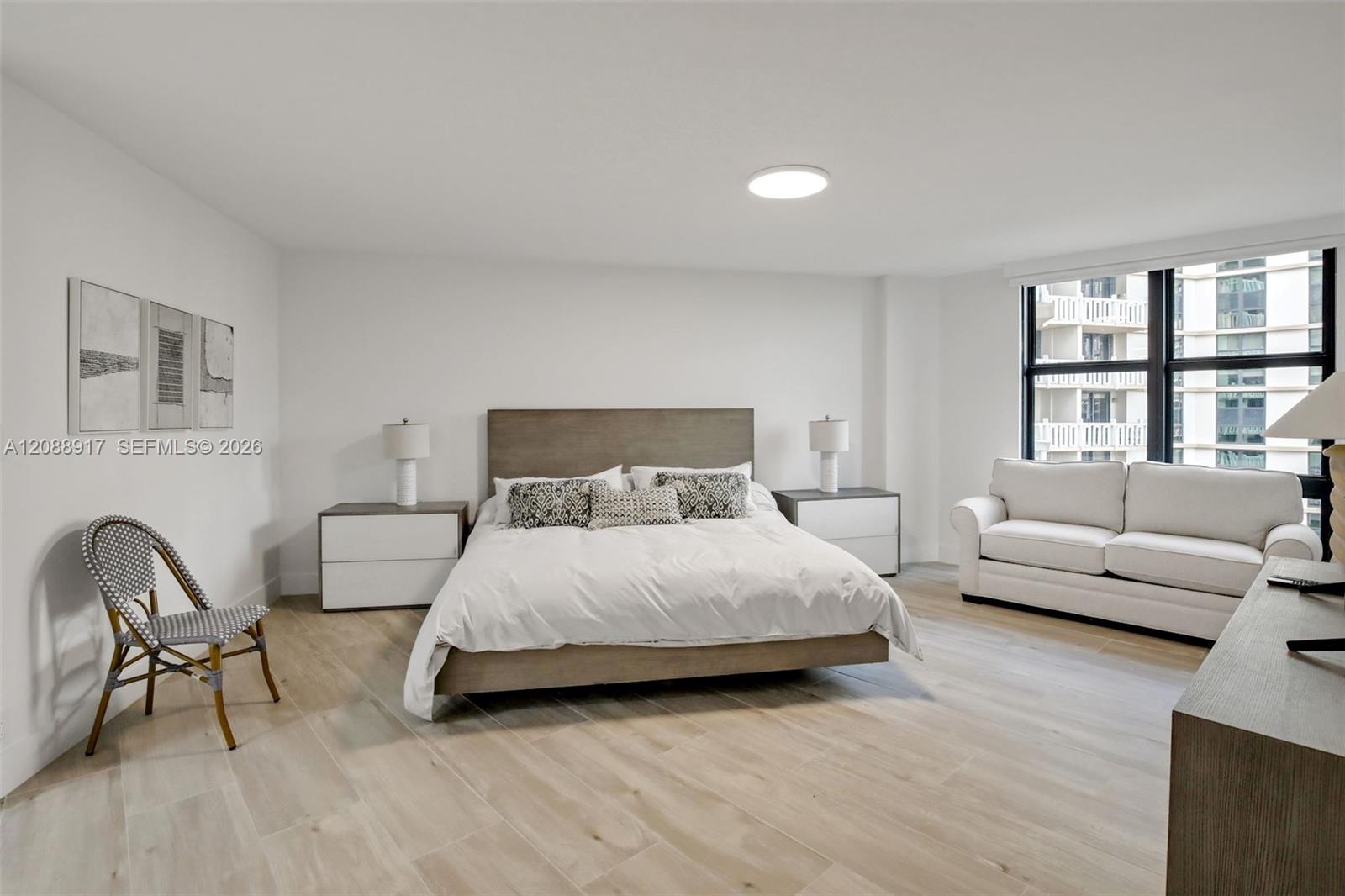 Bright bedroom with a king bed, white sofa, and large windows overlooking a neighboring high-rise.