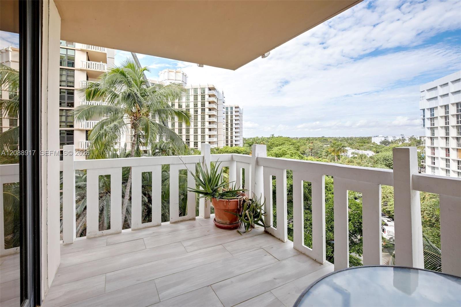 Condominium balcony with tiled flooring, potted plants, and views of palm trees and neighboring buildings.