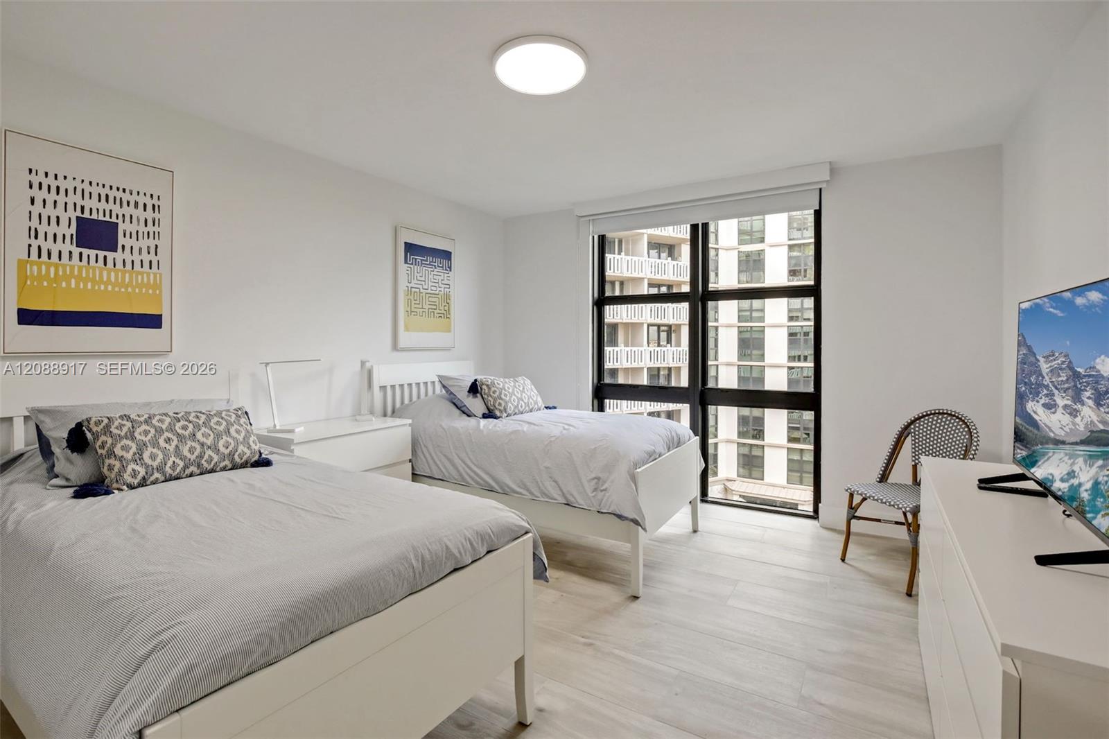 Bedroom with two white beds and large windows overlooking neighboring high-rise buildings.