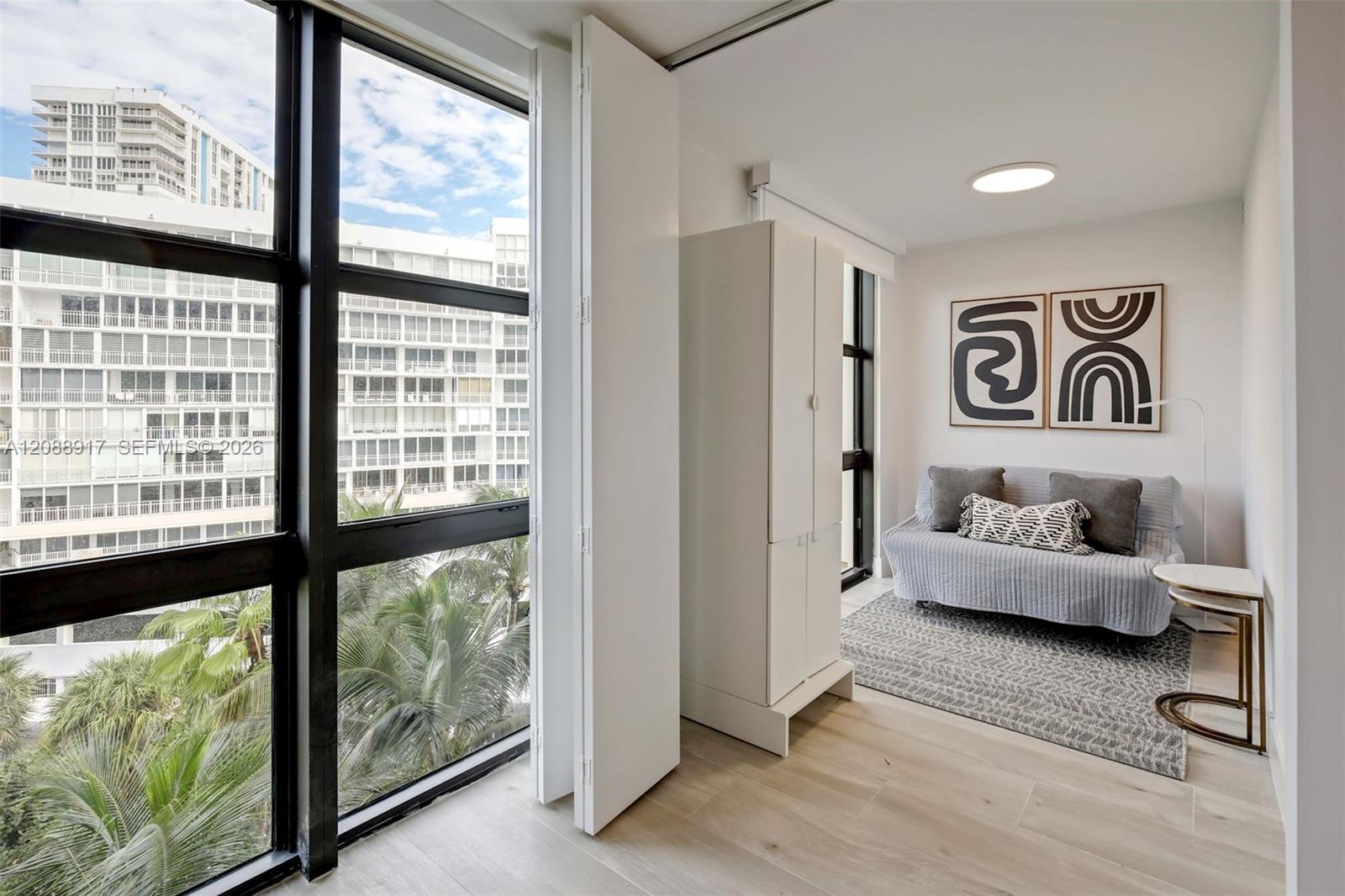 Bright bedroom with a gray daybed, built-in wardrobe, and large windows overlooking neighboring buildings.