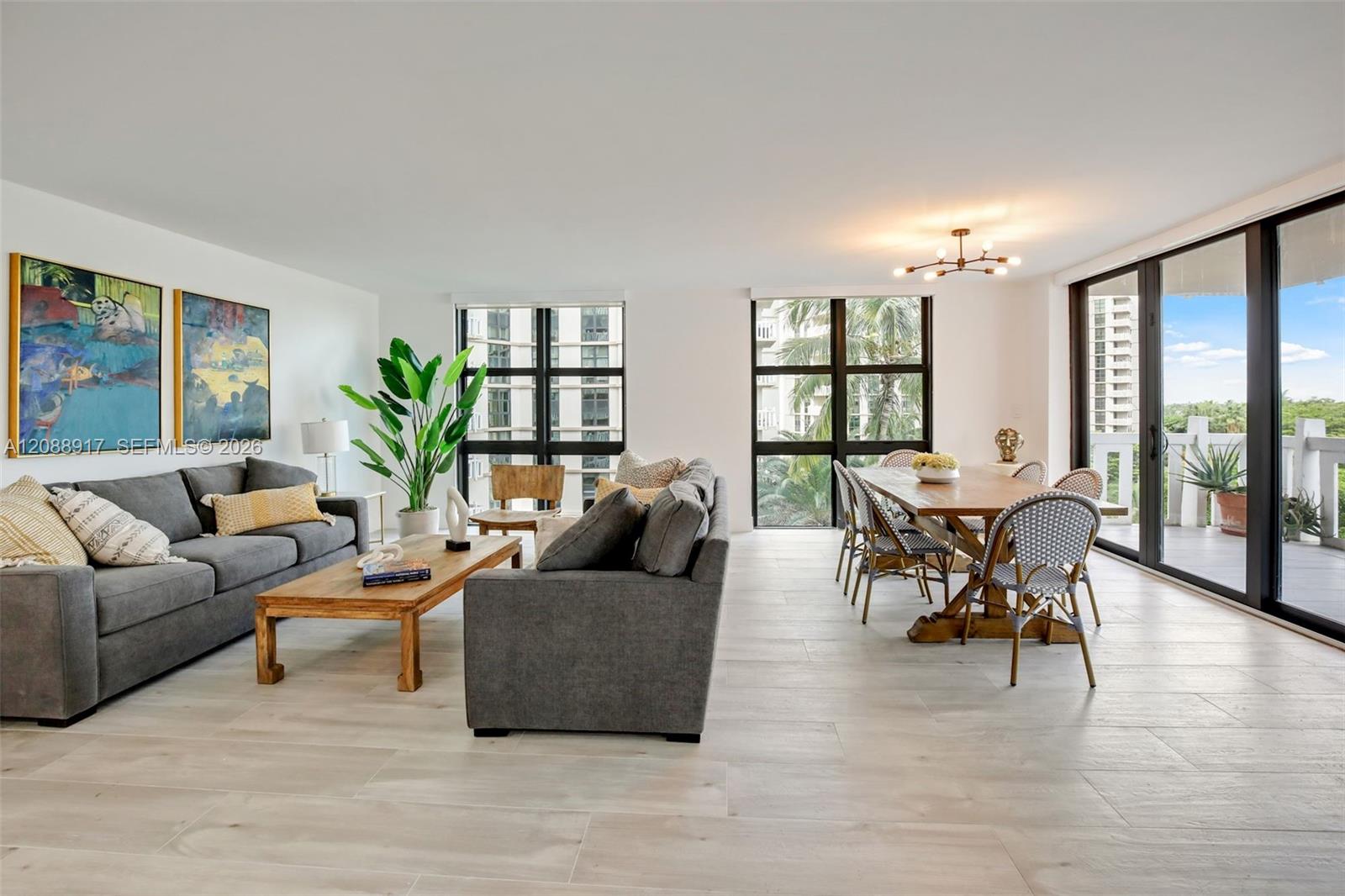Open-plan living and dining room with gray sofas, a wood table, and expansive windows overlooking palm trees.
