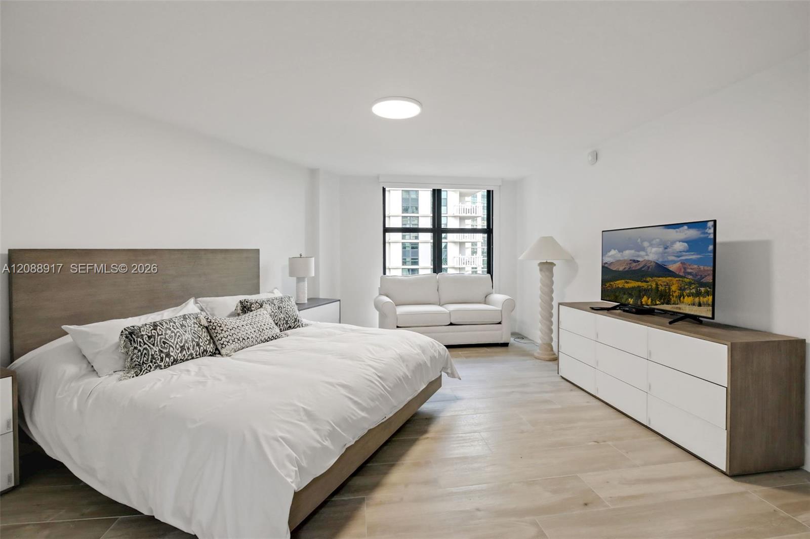 Bedroom with a large bed, patterned pillows, loveseat by the window, and television on a dresser.