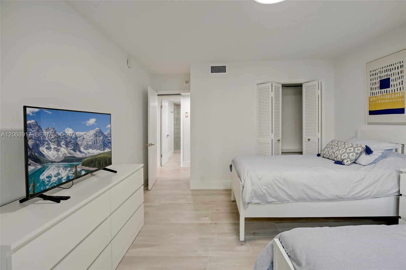 Furnished bedroom with a white bed, TV and dresser, and an open doorway to the bathroom.