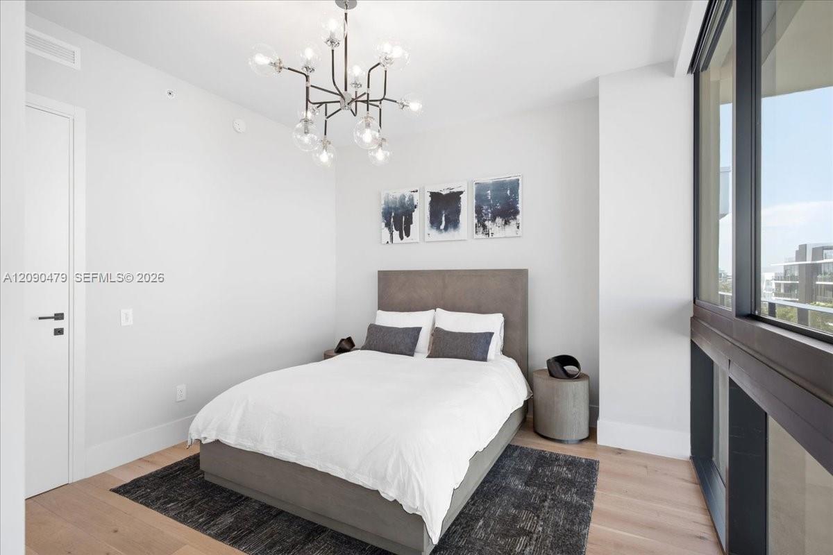 Staged bedroom with a white bed, glass chandelier, and floor-to-ceiling windows overlooking nearby buildings.
