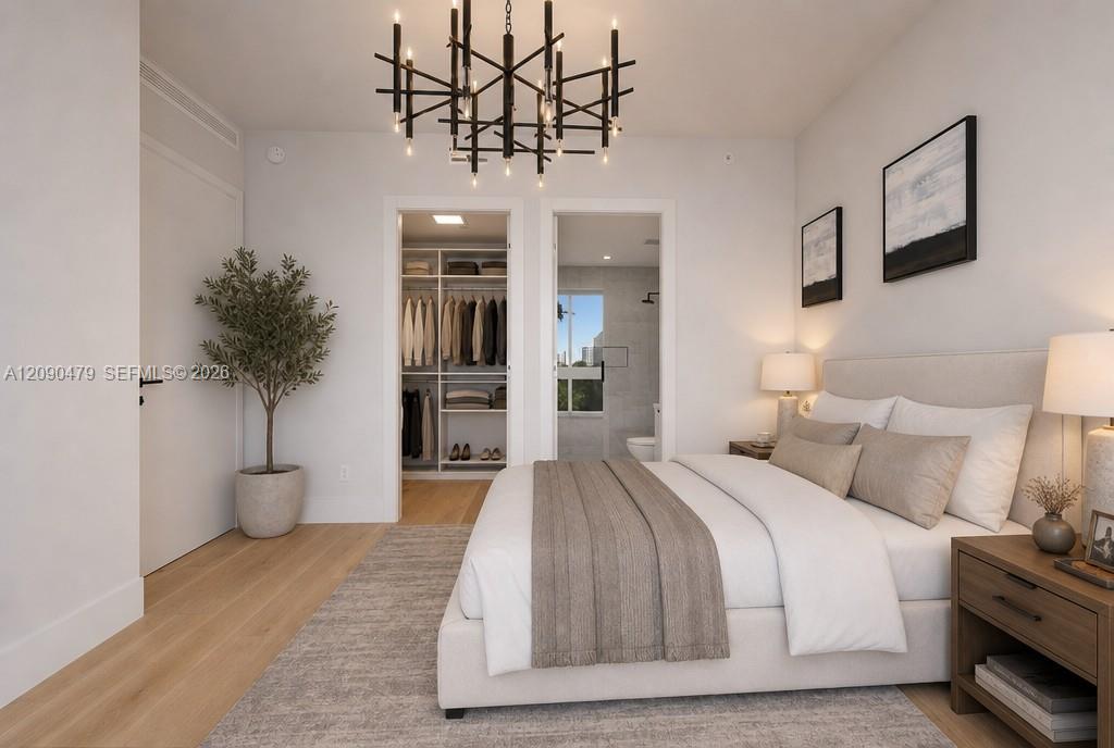 Staged bedroom with an upholstered bed, branching chandelier, open closet, and ensuite bathroom doorway.