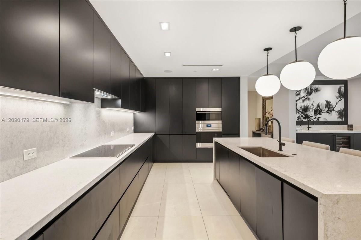 Modern kitchen with dark cabinetry, pale stone counters, a large island, and spherical pendant lights.