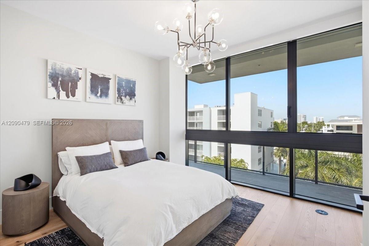Staged bedroom with a white bed and floor-to-ceiling windows overlooking nearby buildings and palms.