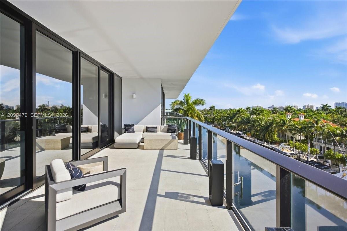 Furnished condo balcony with glass railing, lounge seating, and a sunny view over palms and city buildings.