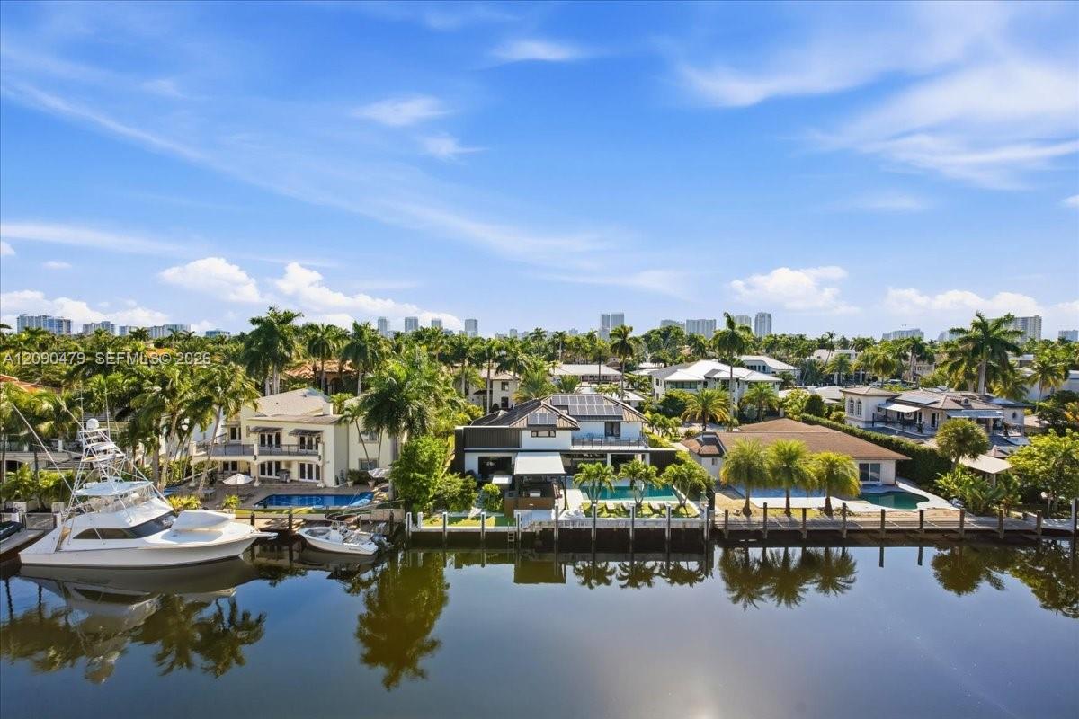 Waterfront canal with docked boats, palm-lined homes, and distant high-rise buildings.