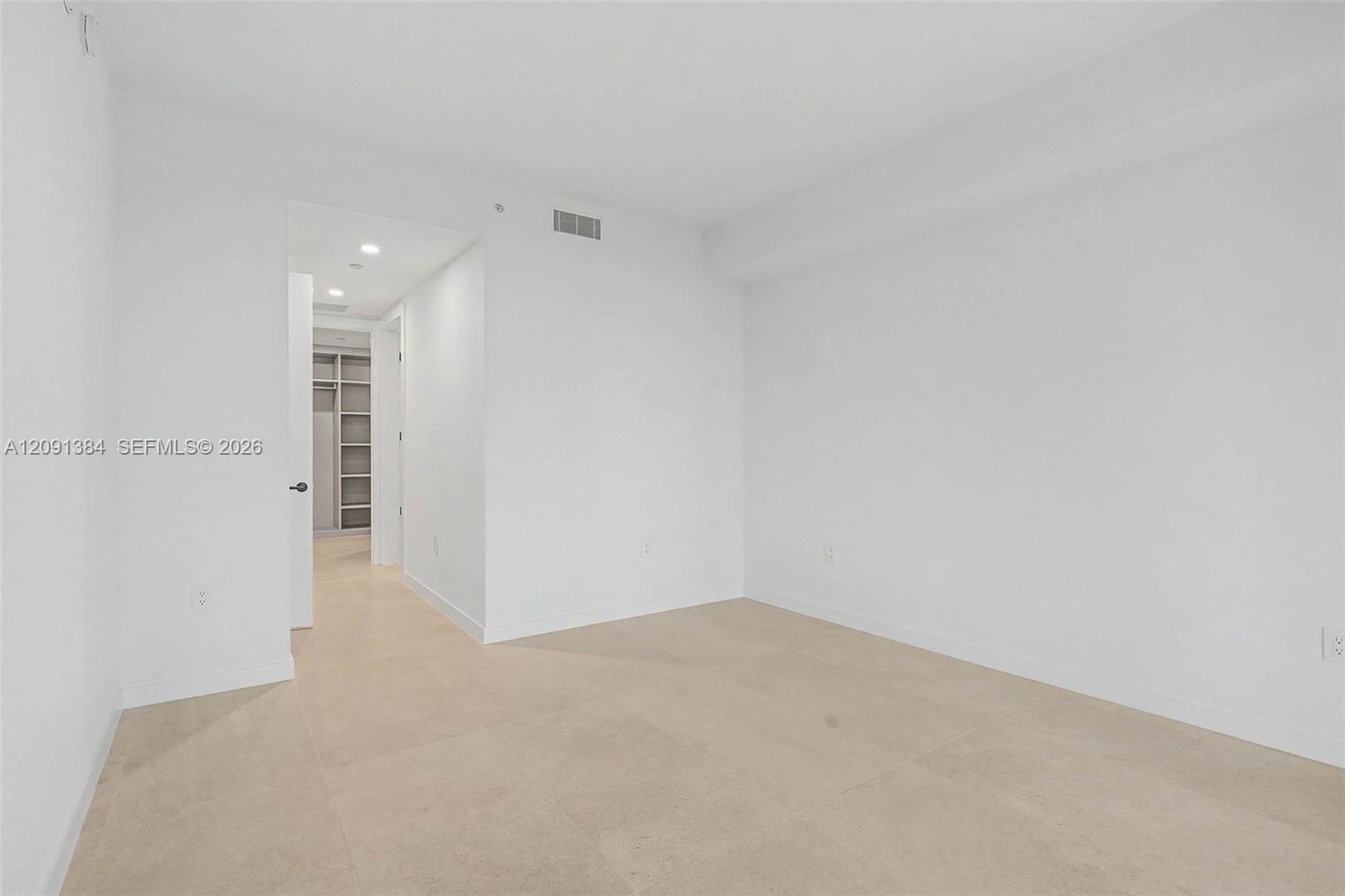 Vacant bedroom with beige tile floors and a doorway to a hall with closet shelving.