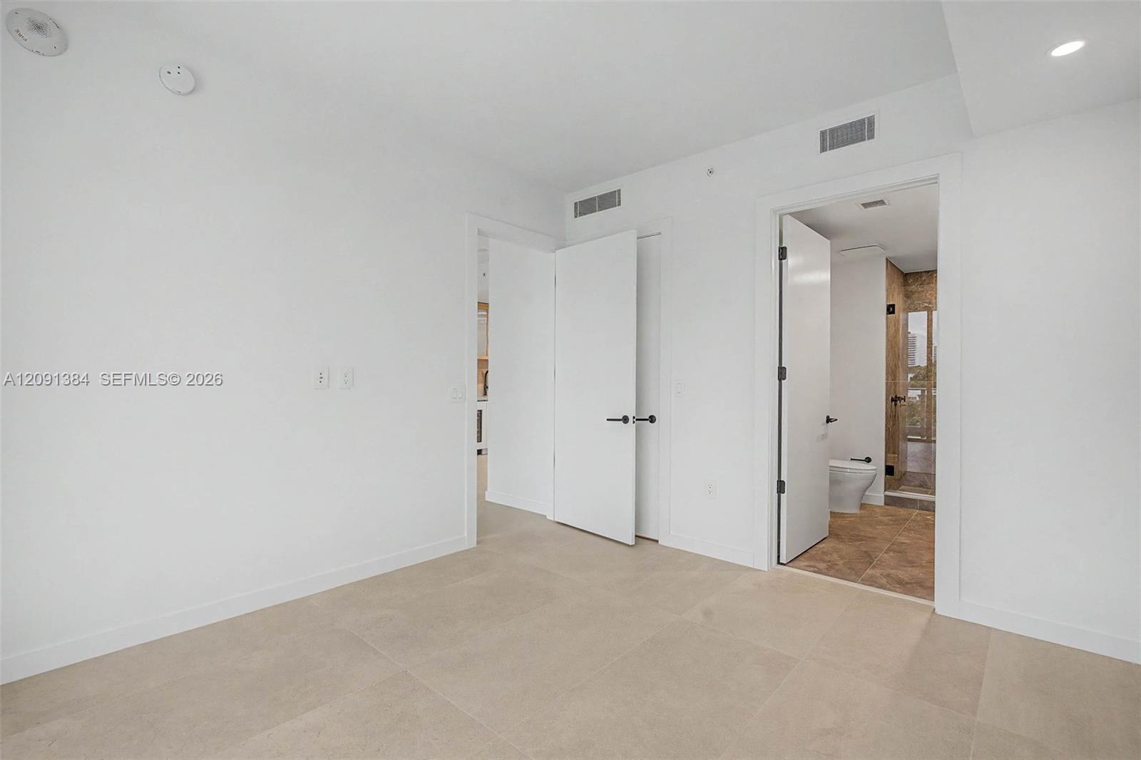 Unfurnished bedroom with beige tile floors and open doors to a bathroom.