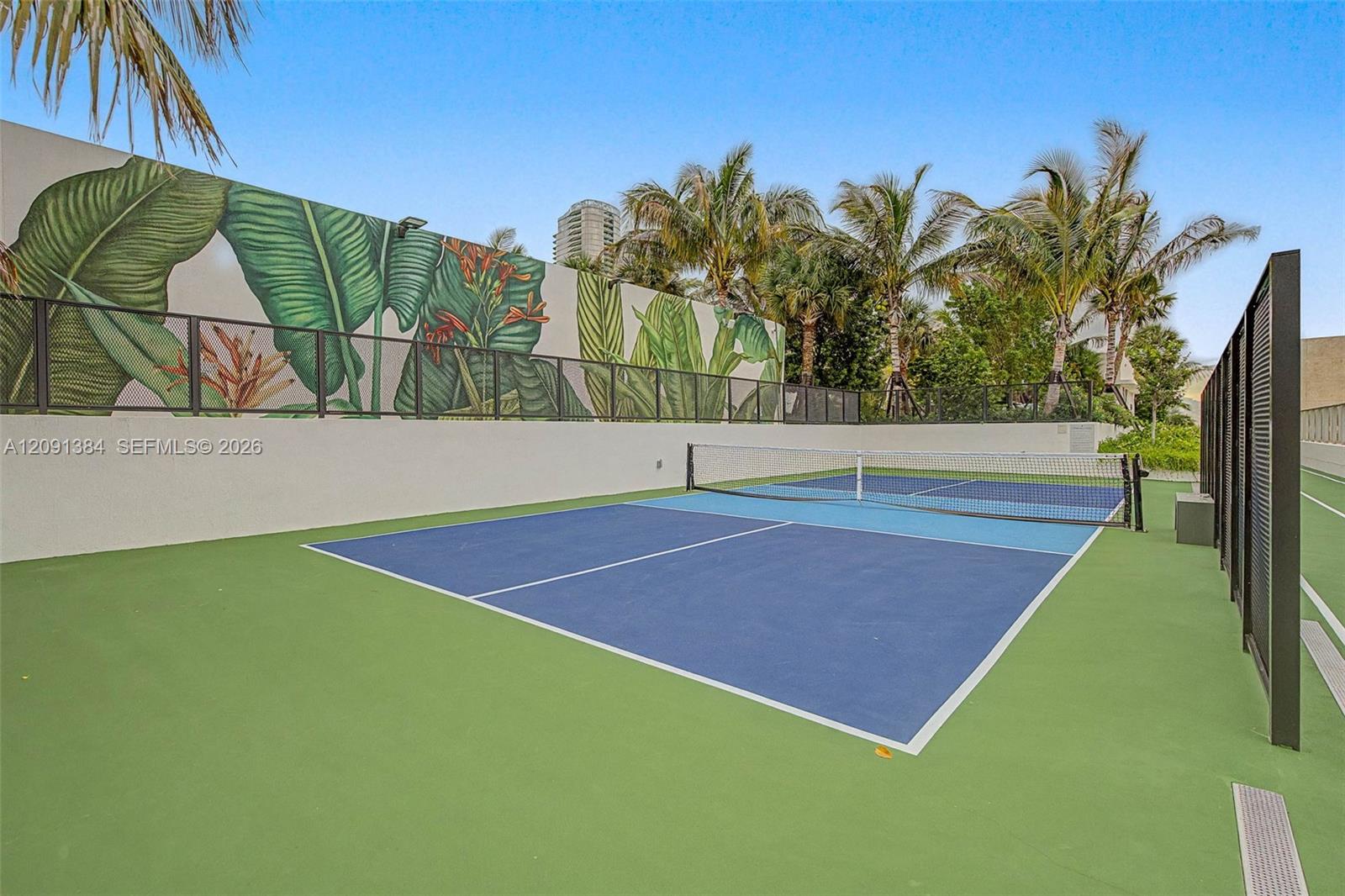 Outdoor pickleball court with a blue playing surface, net, palm trees, and a tropical mural.