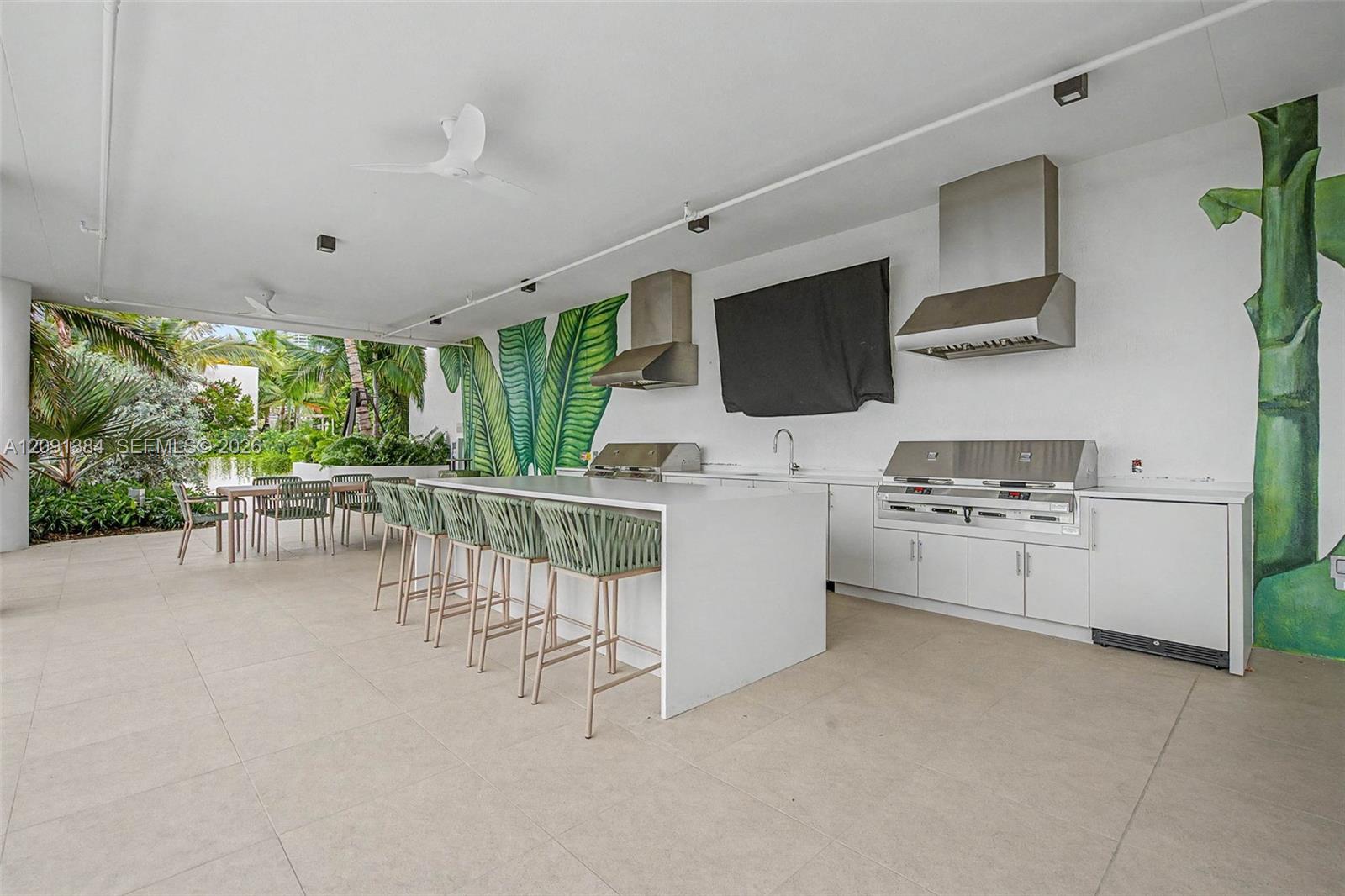 Covered outdoor kitchen with a long bar island, stainless steel grills, and tropical plants.