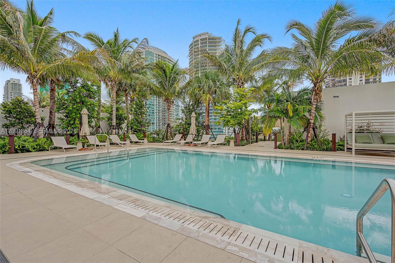 Outdoor swimming pool bordered by palm trees and loungers, with high-rise buildings behind.